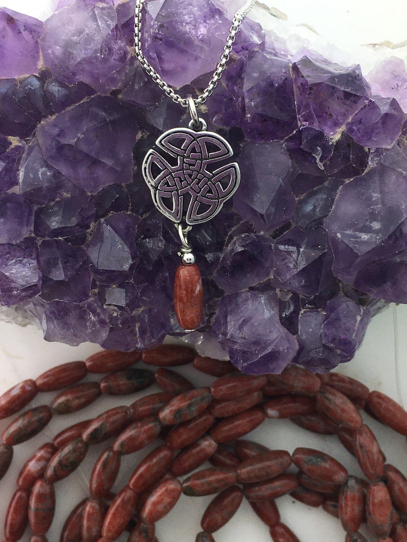 May include: A silver Celtic knot pendant with a purple enamel design, hanging from a silver chain. A red gemstone bead dangles from the pendant.