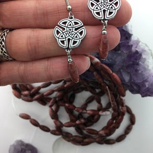 May include: Silver Celtic knot earrings with red jasper stone dangles. The earrings are on a hand with a purple amethyst crystal in the background.