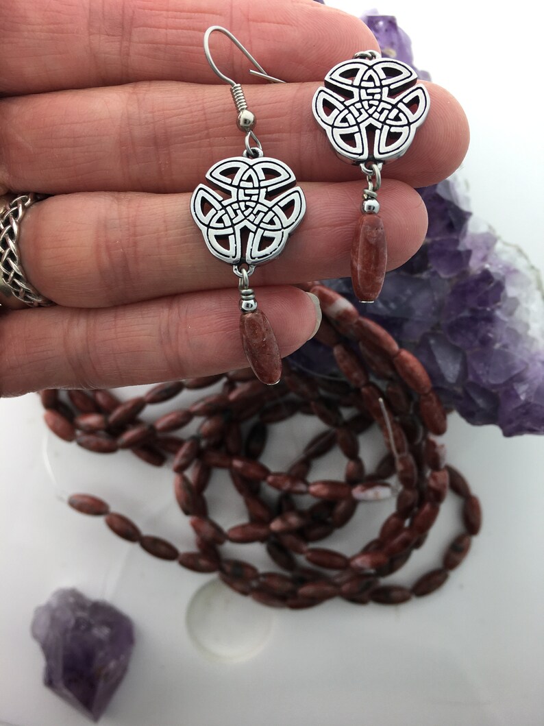 May include: Silver Celtic knot earrings with red jasper stone dangles. The earrings are on a hand with a purple amethyst crystal in the background.