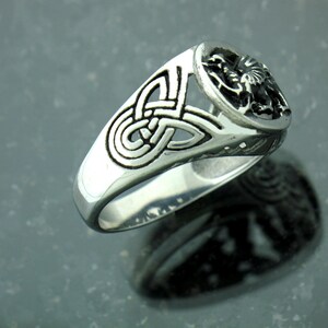 May include: A silver ring with a Celtic knot design and a raised circular design on the top. The circular design features a stylized animal figure.
