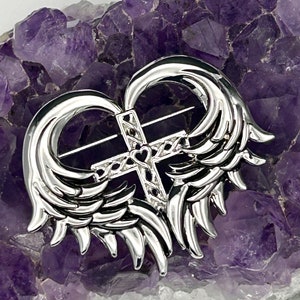 May include: A silver-toned brooch in the shape of a heart with angel wings and a cross in the center. The cross has a small heart in the center.