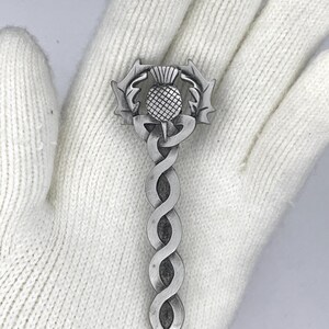 May include: A silver-toned kilt pin featuring a stylized thistle with a Celtic knot design.