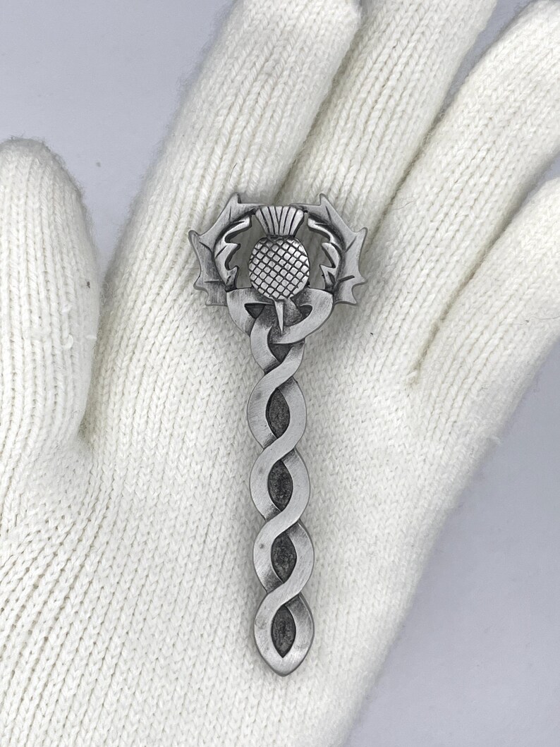 May include: A silver-toned kilt pin featuring a stylized thistle with a Celtic knot design.