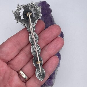 May include: A silver-toned brooch with a detailed design of a stylized flower. The brooch has a pin back and is held in a hand.