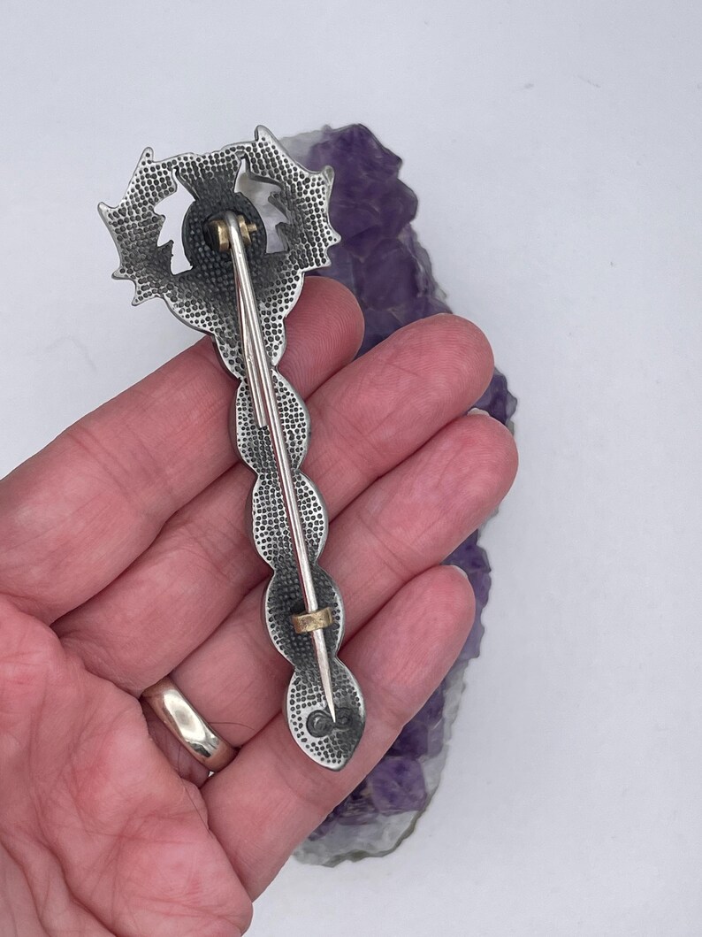 May include: A silver-toned brooch with a detailed design of a stylized flower. The brooch has a pin back and is held in a hand.