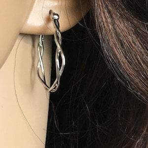 May include: A silver-colored, twisted hoop earring is shown on a person's ear. The earring has a woven design, with the metal appearing shiny and reflective. The earring is a classic jewelry piece, suitable for everyday wear or special occasions.