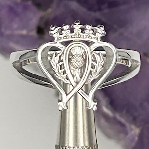 K&ouml;nnte beinhalten: Ein silberner Ring mit einem keltischen Knotendesign, das einen Distel und eine Krone zeigt. Der Ring befindet sich auf einem silbernen St&auml;nder.