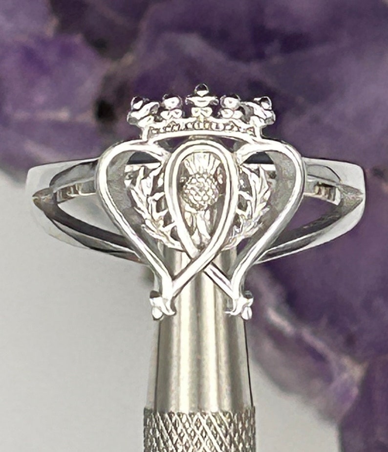 K&ouml;nnte beinhalten: Ein silberner Ring mit einem keltischen Knotendesign, das einen Distel und eine Krone zeigt. Der Ring befindet sich auf einem silbernen St&auml;nder.