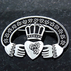 Könnte beinhalten: Silberne Claddagh-Brosche mit einem Herzen, Händen und einer Krone. Das Herz ist mit Kleeblättern verziert. Die Krone ist schwarz.