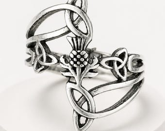 May include: A silver-toned metal ring featuring a central thistle motif surrounded by intricate Celtic knotwork patterns. The design incorporates oxidized detailing within the recessed areas to highlight the sculptural metalwork of the band.