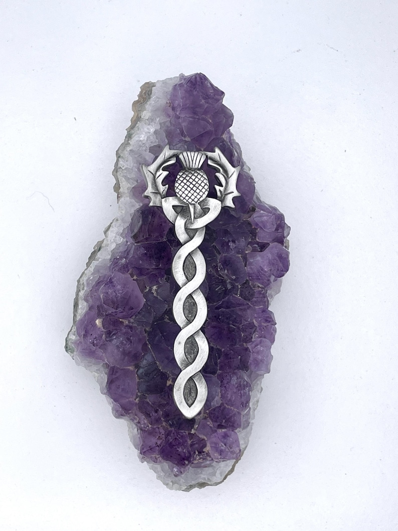 May include: A silver Celtic knot pendant with a thistle design, displayed on a bed of amethyst crystals.