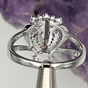 K&ouml;nnte beinhalten: Ein silberner Ring mit einem herzf&ouml;rmigen Design und einer Reihe kleiner Steine auf der Oberseite. Der Ring befindet sich auf einem silbernen St&auml;nder.
