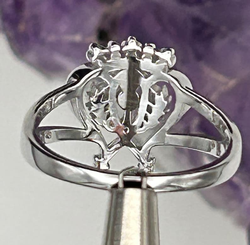 K&ouml;nnte beinhalten: Ein silberner Ring mit einem herzf&ouml;rmigen Design und einer Reihe kleiner Steine auf der Oberseite. Der Ring befindet sich auf einem silbernen St&auml;nder.