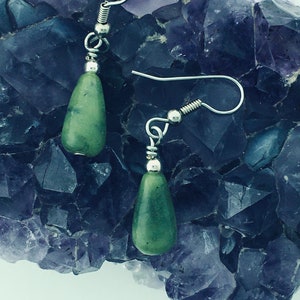 May include: A pair of silver dangle earrings with green gemstone drops. The earrings are displayed on a bed of purple amethyst crystals.