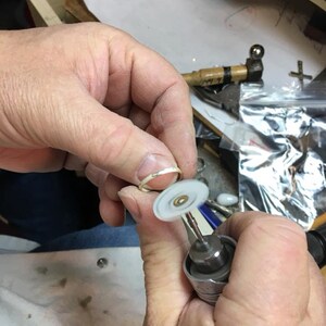 May include: A person is using a small, handheld rotary tool with a white polishing wheel to polish a silver ring.