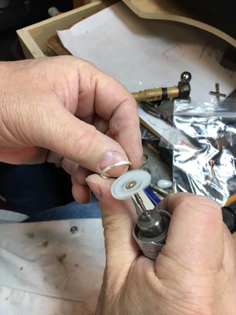May include: A person is using a small, handheld rotary tool with a white polishing wheel to polish a silver ring.