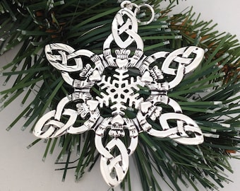 May include: A silver-toned metal ornament shaped like a snowflake, featuring six repeating Claddagh motifs with hands, hearts, and crowns, interconnected by intricate Celtic knotwork patterns. A small circular loop is attached at the top.