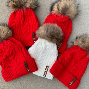 May include: Four red and white knit winter hats with faux fur pom poms. Each hat has a personalized leather patch with a name on it. The names are: Liz, Kassie, Mrs. Farmer, and Giz.
