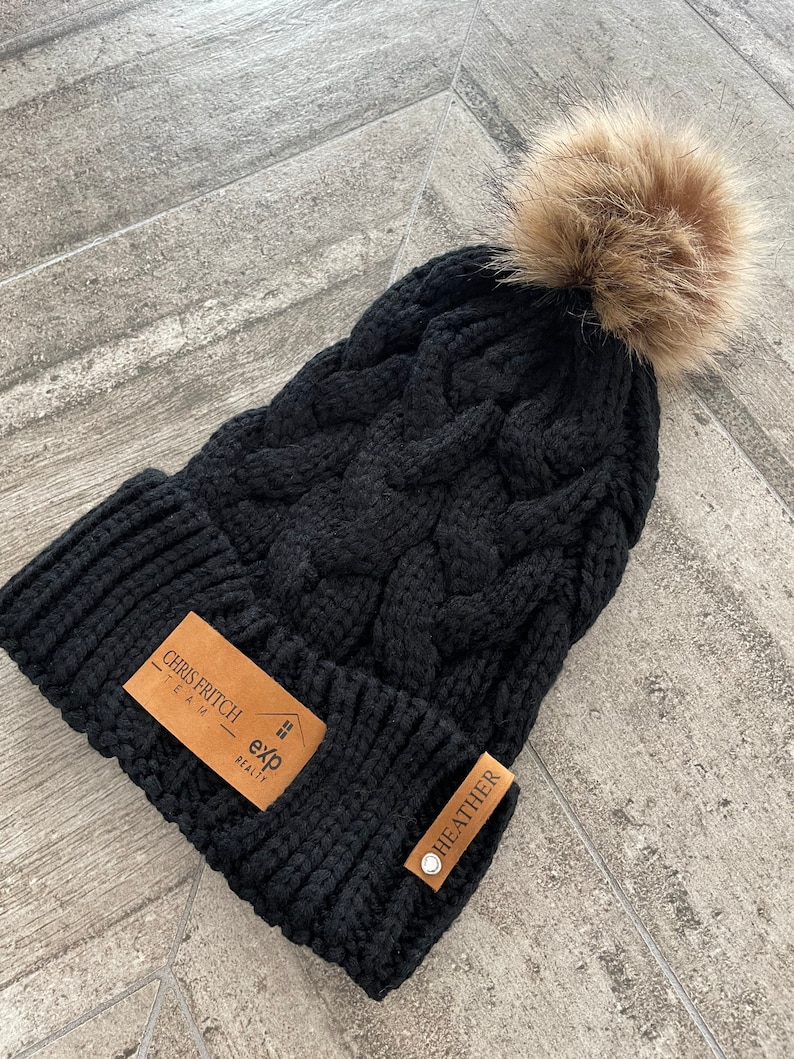 May include: Black cable knit beanie with a brown faux fur pom pom. The beanie has two brown leather tags with the text "Chris Fritsch Team exp Realty" and "Heather" on them.