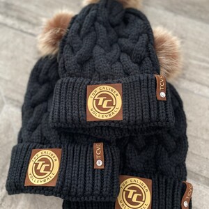 May include: Three black knit winter hats with faux fur pom poms. Each hat has a brown leather patch with a gold circle logo that says "Top Caliber Volleyball" and a brown leather tag that says "TCVC".