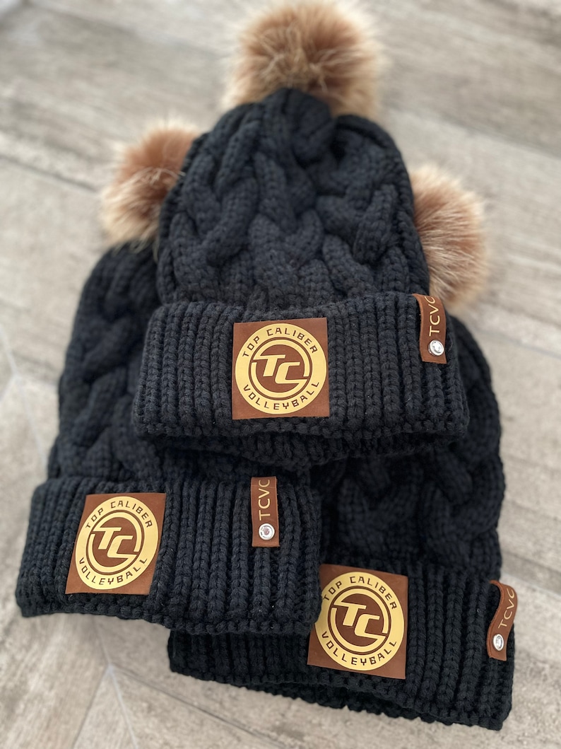 May include: Three black knit winter hats with faux fur pom poms. Each hat has a brown leather patch with a gold circle logo that says "Top Caliber Volleyball" and a brown leather tag that says "TCVC".