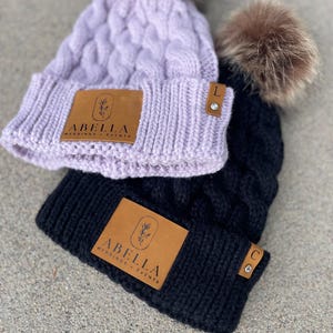 May include: Two knitted beanies with faux fur pom poms. Both beanies have a brown leather patch with the text "Abella Weddings + Events". The beanie on the left is lavender and has a letter "L" charm. The beanie on the right is black and has a letter "C" charm.
