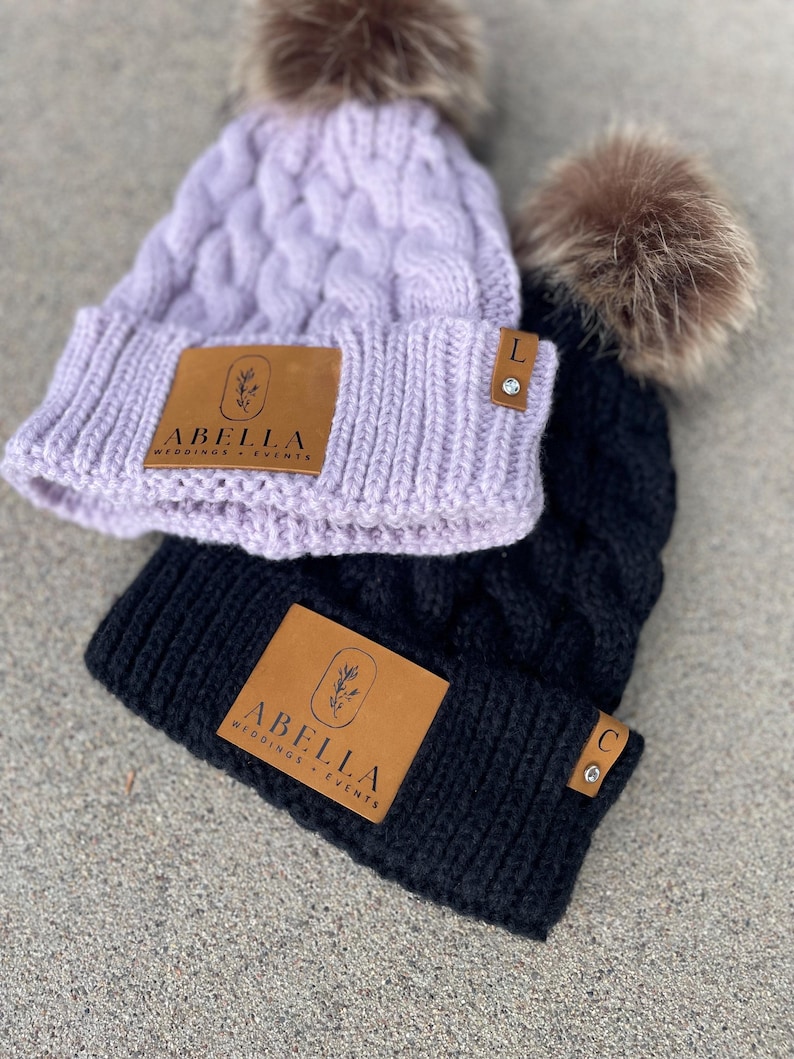May include: Two knitted beanies with faux fur pom poms. Both beanies have a brown leather patch with the text "Abella Weddings + Events". The beanie on the left is lavender and has a letter "L" charm. The beanie on the right is black and has a letter "C" charm.