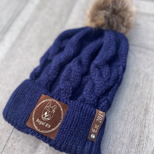 May include: A navy blue knitted beanie with a brown faux fur pom pom. The beanie has a brown leather patch with a dog silhouette and the text "Regal K9" and a brown leather tag with the text "Est. 2019".