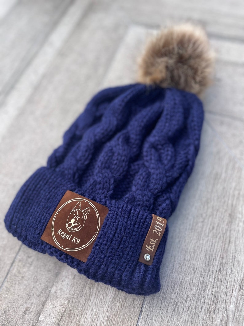 May include: A navy blue knitted beanie with a brown faux fur pom pom. The beanie has a brown leather patch with a dog silhouette and the text "Regal K9" and a brown leather tag with the text "Est. 2019".