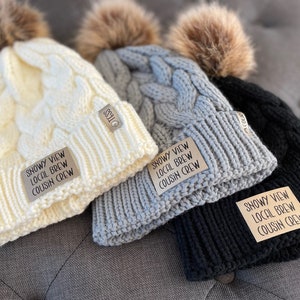 May include: Three knitted winter hats with faux fur pom poms. Each hat has a brown leather patch with the text "Snowy View Local Brew Cousin Crew".