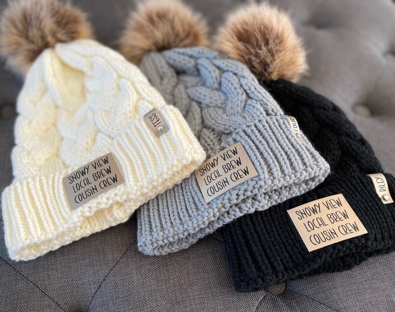 May include: Three knitted winter hats with faux fur pom poms. Each hat has a brown leather patch with the text "Snowy View Local Brew Cousin Crew".
