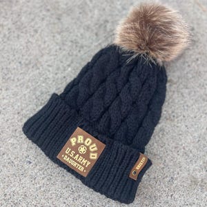May include: Black knitted beanie with a brown faux fur pom pom. The beanie has a brown leather patch with gold lettering that reads "Proud U.S. Army Daughter".