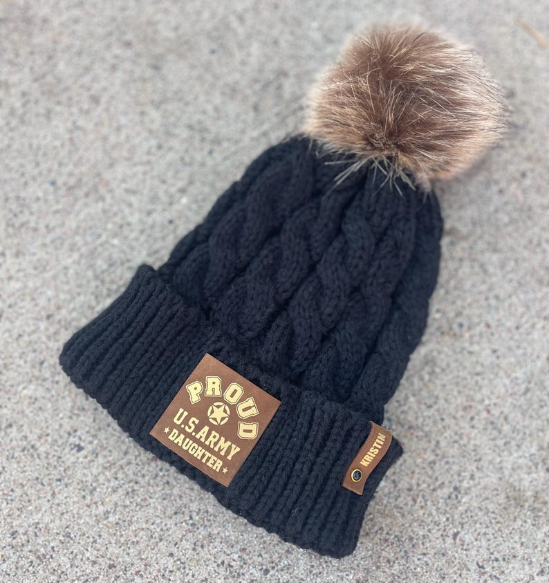 May include: Black knitted beanie with a brown faux fur pom pom. The beanie has a brown leather patch with gold lettering that reads "Proud U.S. Army Daughter".