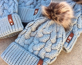 May include: Three gray knitted hats with faux fur pom poms. The hats have leather patches with the letters A, G, and L on them. The hats are on a light brown surface.