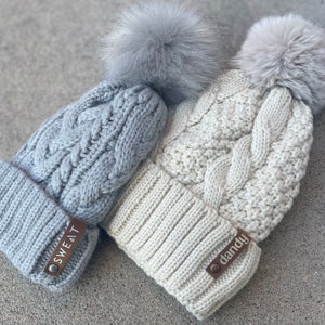 May include: Two cable-knit beanies with faux fur pom-poms. One is light gray with a leather tag that reads "SWEAT", and the other is off-white with a tag that reads "dandy". The beanies are made of a soft, textured material.