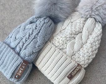 May include: Two cable-knit beanies with faux fur pom-poms. One is light gray with a leather tag that reads "SWEAT", and the other is off-white with a tag that reads "dandy". The beanies are made of a soft, textured material.