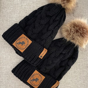 May include: Two black knit hats with faux fur pom poms. Each hat has a brown leather patch with the text "Belle Mountain" and a horse silhouette. The patches also have the text "Madilyn" and "Reagan" on them.