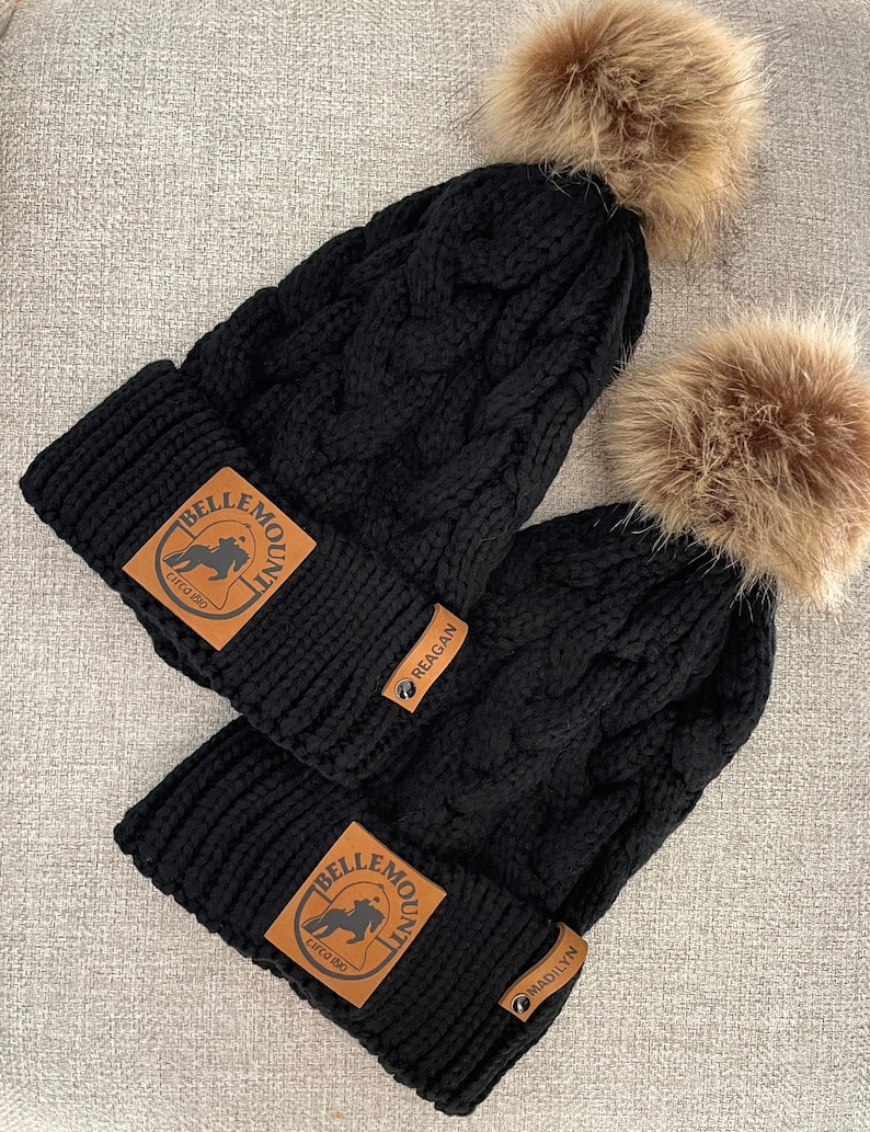 May include: Two black knit hats with faux fur pom poms. Each hat has a brown leather patch with the text "Belle Mountain" and a horse silhouette. The patches also have the text "Madilyn" and "Reagan" on them.