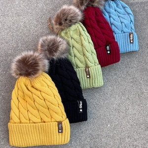 May include: Five knitted winter hats with faux fur pom poms in various colors, including yellow, black, green, red, and blue. Each hat has a small leather tag with the word "Filly" on it.