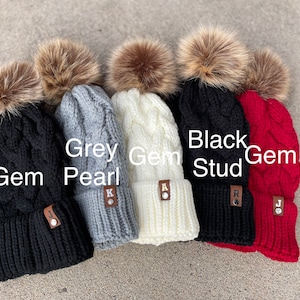 May include: Five knitted winter hats with faux fur pom poms in black, grey, white, and red. The hats have a cable knit pattern and a leather patch with a letter on each.