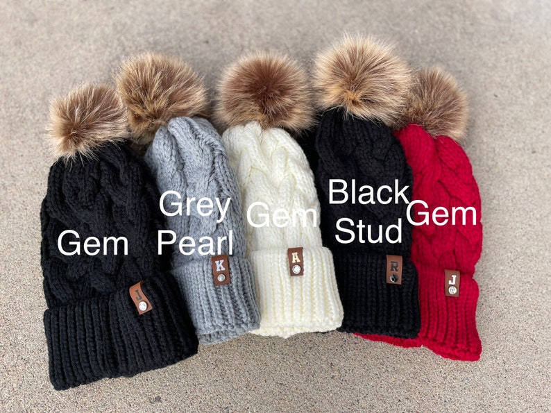 May include: Five knitted winter hats with faux fur pom poms in black, grey, white, and red. The hats have a cable knit pattern and a leather patch with a letter on each.