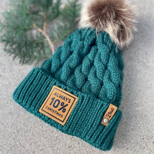 May include: A dark green knitted beanie with a brown faux fur pom pom. The beanie has a brown leather patch with the text "ALWAYS 10% CHRISTMAS" and a small brown leather tag with the text "Linds".