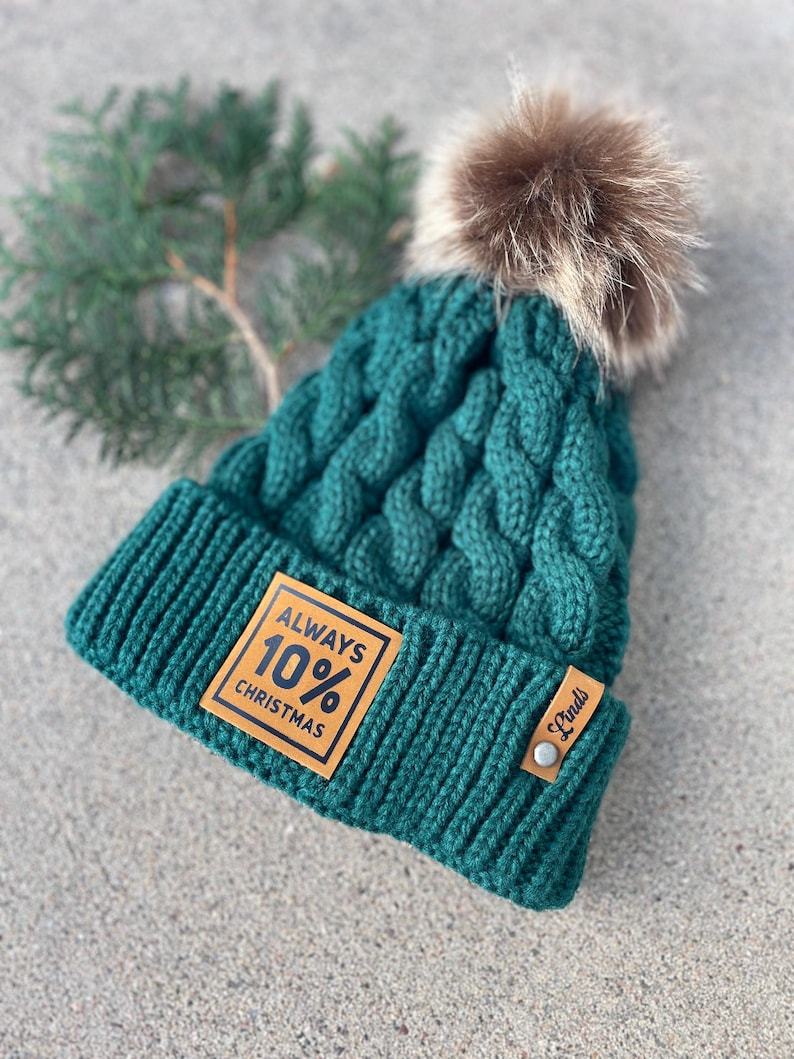 May include: A dark green knitted beanie with a brown faux fur pom pom. The beanie has a brown leather patch with the text "ALWAYS 10% CHRISTMAS" and a small brown leather tag with the text "Linds".