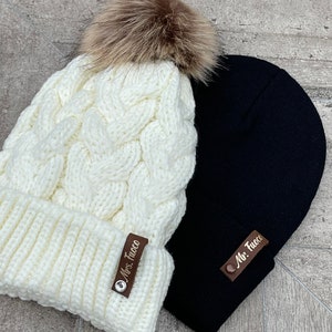 May include: Two knit winter hats, one white with a brown faux fur pom pom and the other black. Both hats have a brown leather patch with the text "Mrs. Tuco" and "Mr. Tuco" respectively.
