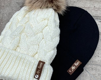 May include: Two knit winter hats, one white with a brown faux fur pom pom and the other black. Both hats have a brown leather patch with the text "Mrs. Tuco" and "Mr. Tuco" respectively.