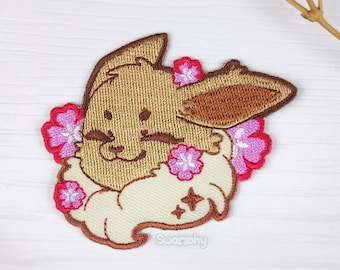 May include: Embroidered patch featuring a brown Eevee with pink flowers around its head. The Eevee is smiling and has a light brown fluffy tail. The patch is on a white background.
