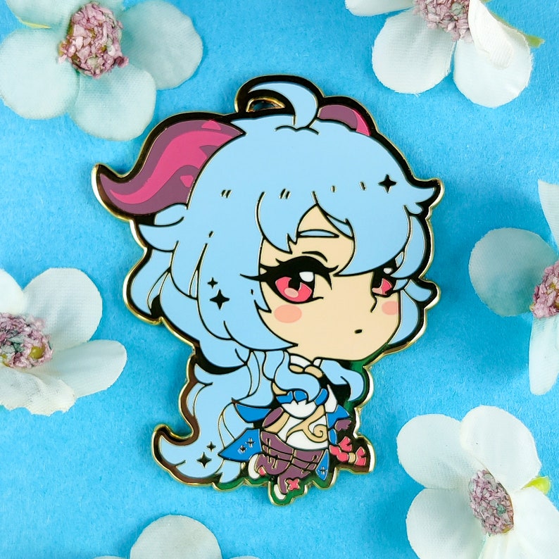 May include: A blue and pink enamel pin featuring a cartoon character with long blue hair, pink horns, and a gold outline. The character is wearing a white and gold outfit and has a pink blush on their cheeks.