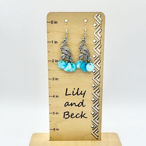 May include: Silver-toned peacock earrings with turquoise and white accents. The earrings are displayed on a wooden stand that reads "Lily and Beck". The earrings are approximately 2 inches long.