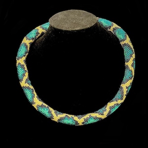 Może przedstawiać: Naszyjnik z koralików o geometrycznym wzorze. Naszyjnik ma powtarzający się wzór z turkusowych, czarnych i żółtych koralików. Naszyjnik jest prezentowany na czarnym biuście jubilerskim na czarnym tle.