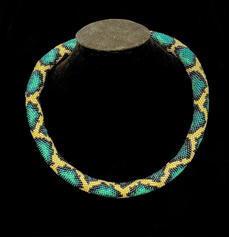 K&ouml;nnte beinhalten: Eine Halskette mit Perlen und einem geometrischen Muster. Die Halskette hat ein sich wiederholendes Design aus t&uuml;rkis, schwarzen und gelben Perlen. Die Halskette wird auf einer schwarzen Schmuckb&uuml;ste vor schwarzem Hintergrund pr&auml;sentiert.